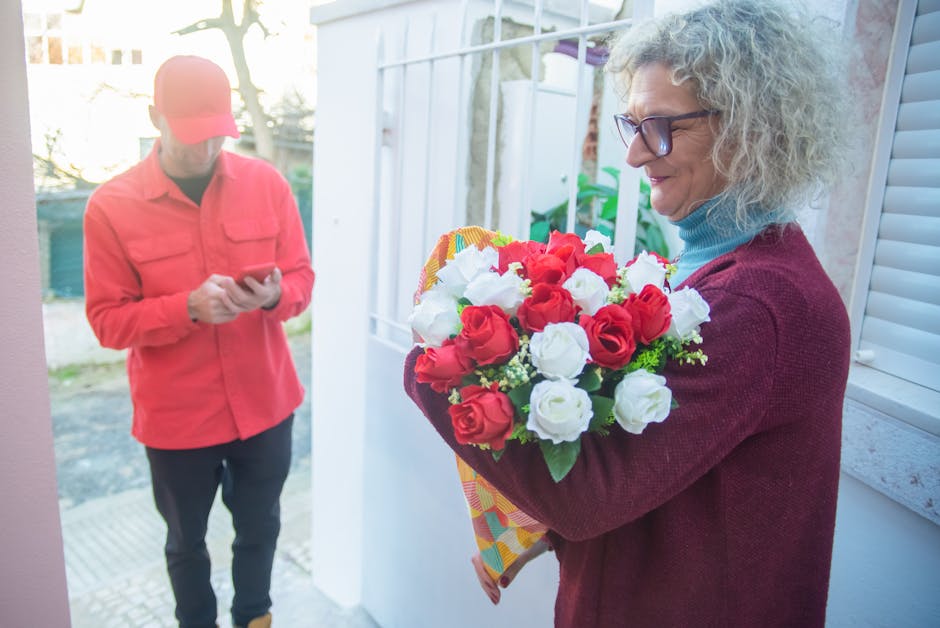 A woman with curly gray hair and glasses holds a vibrant floral bouquet featuring red and white roses, arranged in a compact, rounded style, wrapped in colorful patterned paper. She is standing outdoo