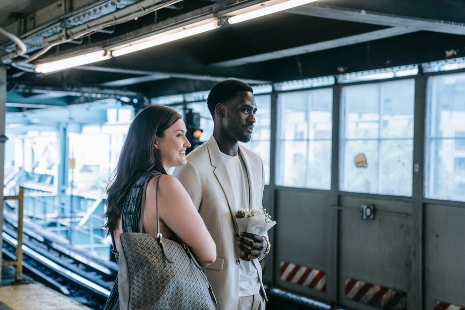 A woman and a man standing on a train or subway platform inside an industrial station with large windows allowing natural light. The woman, smiling and holding a beige handbag, has shoulder-length dar