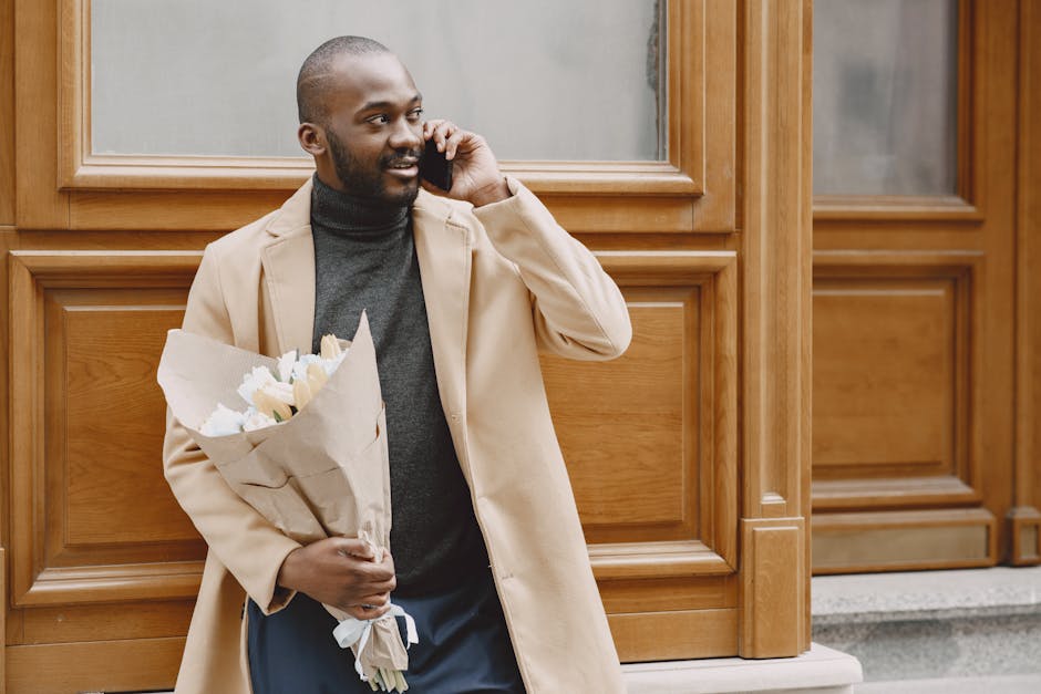 A man with a dark complexion, short hair, and beard, dressed in a beige overcoat and dark turtleneck, is standing outdoors against a wooden-paneled wall. He is holding a bouquet of fresh white and cre