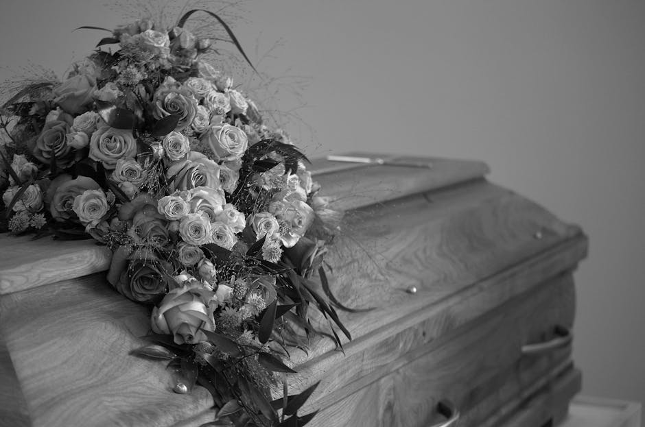 A floral arrangement of fresh roses and other mixed flowers, predominantly in light and dark shades, is placed atop a wooden casket in a funeral setting. The bouquet includes roses with tightly curled