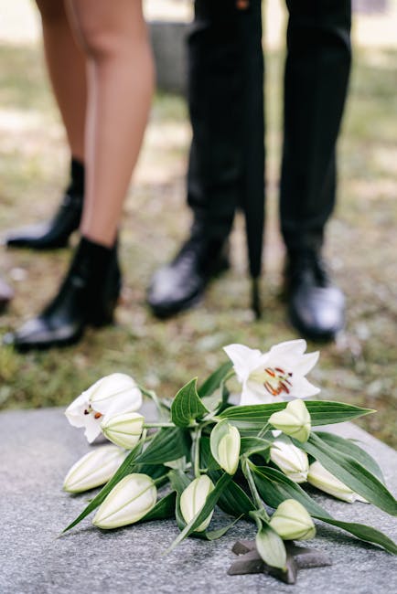 A floral arrangement of white lilies with open blooms and elongated leaves lies on a stone surface, with additional buds and green foliage. In the background, two people stand outdoors on a grassy are