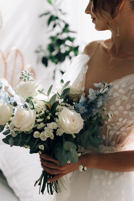 A woman in a white wedding dress holding a bouquet of fresh flowers, including white roses, small white daisies, and blue accents, all arranged with green foliage. The bouquet is wrapped in elegant wh