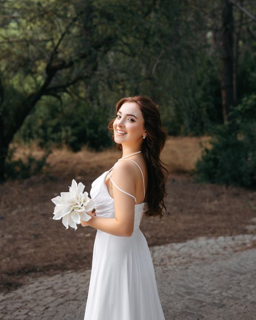 A smiling young woman with long, wavy brown hair wearing a white wedding dress with delicate spaghetti straps, holding a bouquet of large, white, fully bloomed lilies. She stands outdoors in a natural