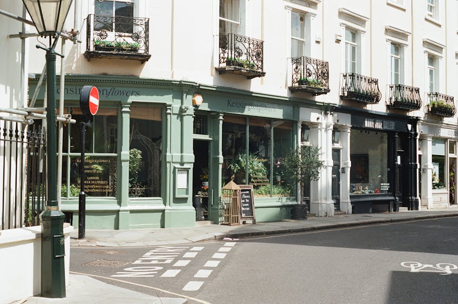 A close-up view of a florist shop on a city street with large glass windows displaying various vibrant flower arrangements. The shop has a soft green exterior with white accents, and a black chalkboar
