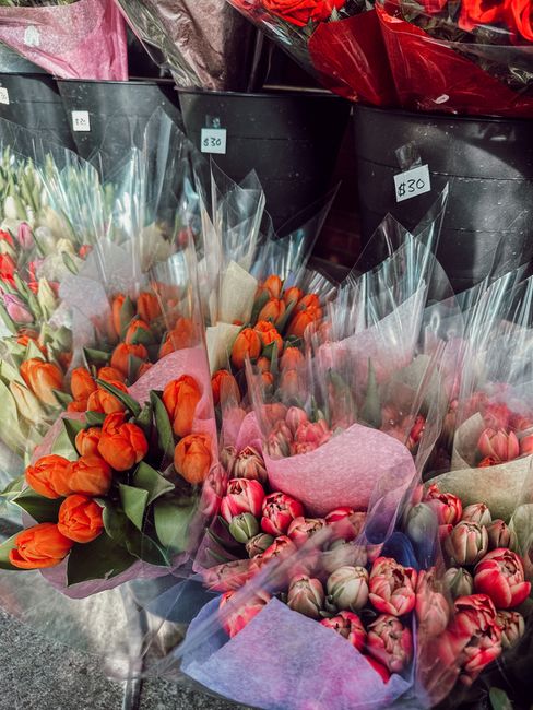 A display of fresh flower bouquets arranged in black containers, featuring tulips and ranunculi in vibrant shades of orange, pink, and red. The tulips have smooth, elongated petals and rich green stem