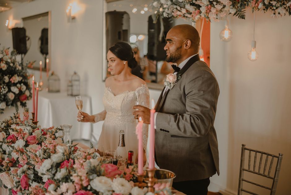 A bride and groom stand at a wedding reception table decorated with a large floral arrangement featuring pink, white, and peach roses, complemented by green foliage. The bride, dressed in an elegant i