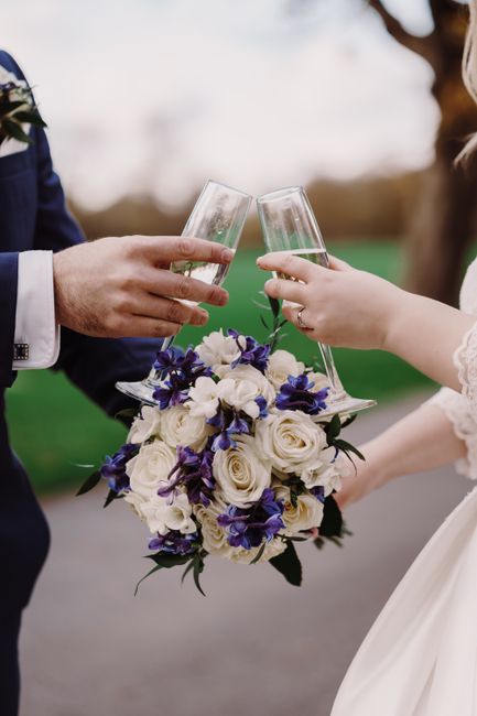 A close-up of a floral bouquet featuring white roses and deep purple irises, held by a bride in a white dress and a groom in a dark suit. The bouquet is arranged with lush green foliage and wrapped in