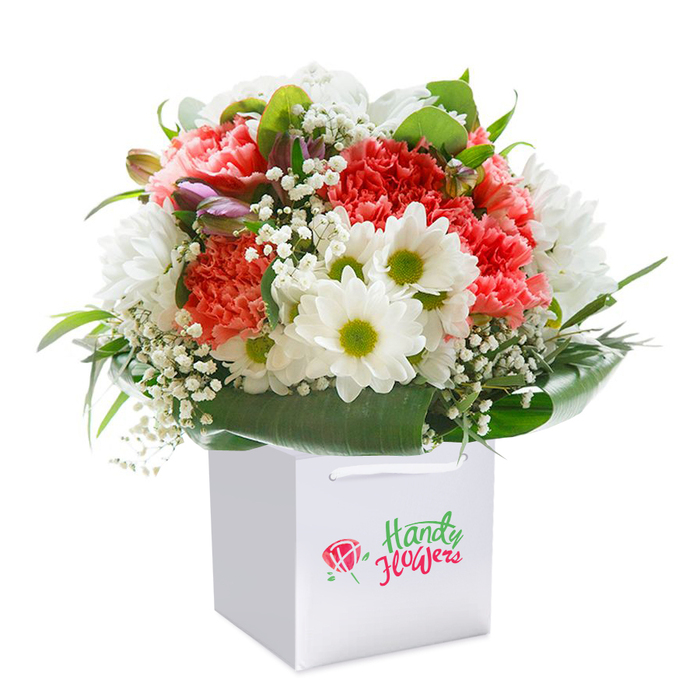 Expertly arranged, this colourful bouquet offers an inviting blend of texture, freshness, and local artistry, reflecting the friendly spirit of Motspur Park. At its heart, coral and soft peach carnations are layered beautifully, their frilled petals giving depth and softness that feels both lively and sophisticated. Bright white daisies-each with a striking lime-tinted centre-nestle beside the carnations, bringing an uplifting brightness reminiscent of a morning stroll through local parks. Gracefully scattered, baby's breath infuses the bouquet with feathery, cloud-like clusters, softening the overall look and drawing the eye toward subtle purple-hued buds tucked thoughtfully on the left. Around the base, large, deep green leaves arc outward, forming a lush, protective collar that strengthens the arrangement and highlights its fullness. Staged upright in a crisp white square gift bag-suitable for easy carrying to Piccadilly Line commutes or doorstep delivery along Motspur Park's tree-lined roads-this bouquet highlights the meticulous care and passion local florists bring to every creation. Soft daylight reveals every detail, from the velvety richness of petals to the vibrant contrast of foliage, offering a sense of generosity and lasting delight. Ideal for an anniversary, just because, or for extending warm wishes to neighbours, this bouquet is a vivid tribute to Motspur Park's floral tradition.